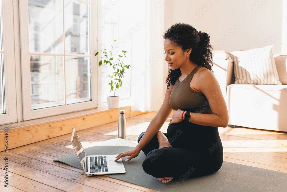 African american pregnant woman looking at laptop screen, searching for online workout tutorial or prenatal yoga sitting on floor on mat in sports wear, wearing smart watch to control heart rate